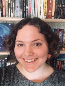 Headshot of a woman with brown, curly hair smiling into the camera. She is wearing red King Arthur earrings, and is standing in front of a bookshelf.